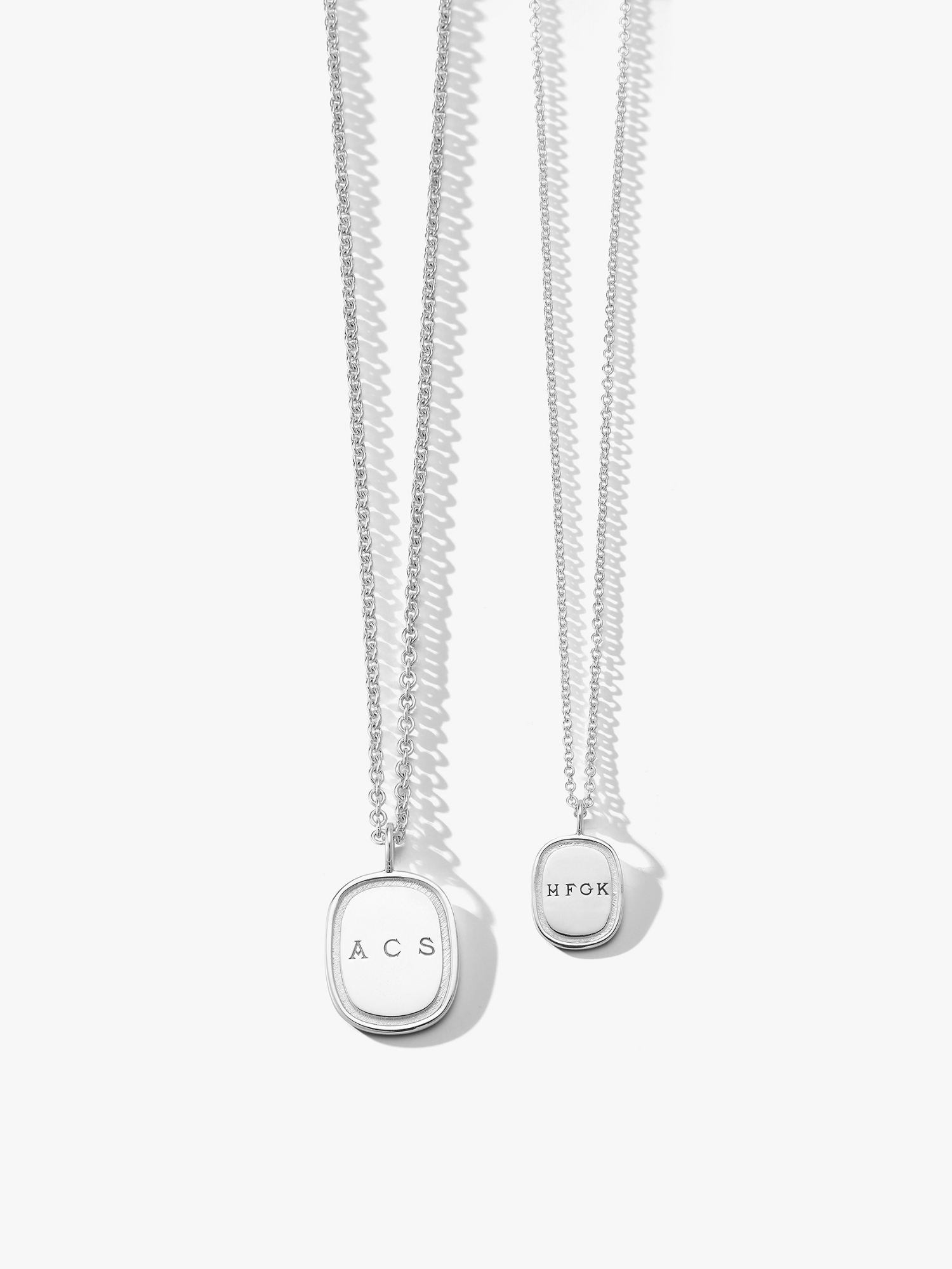 Two silver oblong necklaces. The one on the left is a large oblong necklace engraved with "ACS" and the one on the right is a small silver oblong necklace engraved with."HFGK". Made by Love Billy in NYC.
