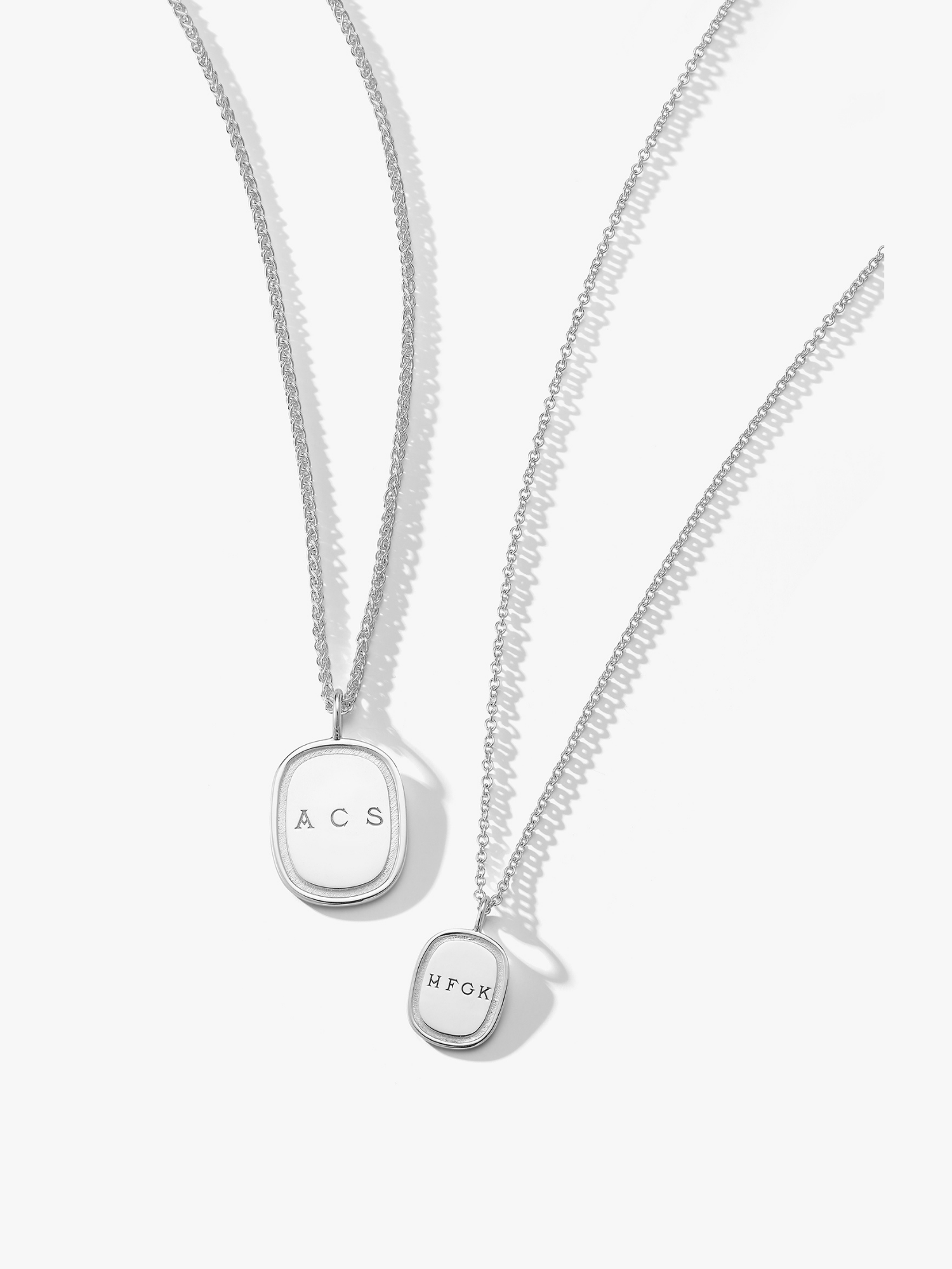 Two silver oblong pendant necklaces. The one on the left is a large oblong pendant engraved with "ACS" on a wheat chain. The one on the right is a small silver oblong pendant on a cable chain engraved with "HFGK". Made by Love Billy.