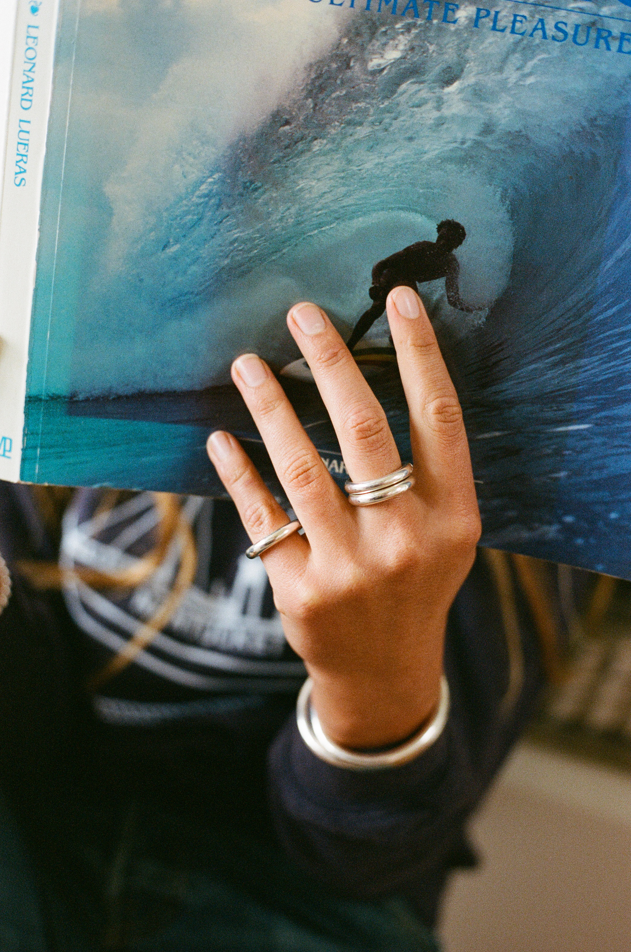 Hand holding a surf book wearing three silver carousel rings. Two are worn on the middle finger and one on the pinky. In the background you can also see a silver carousel cuff. Made by love billy in NYC.