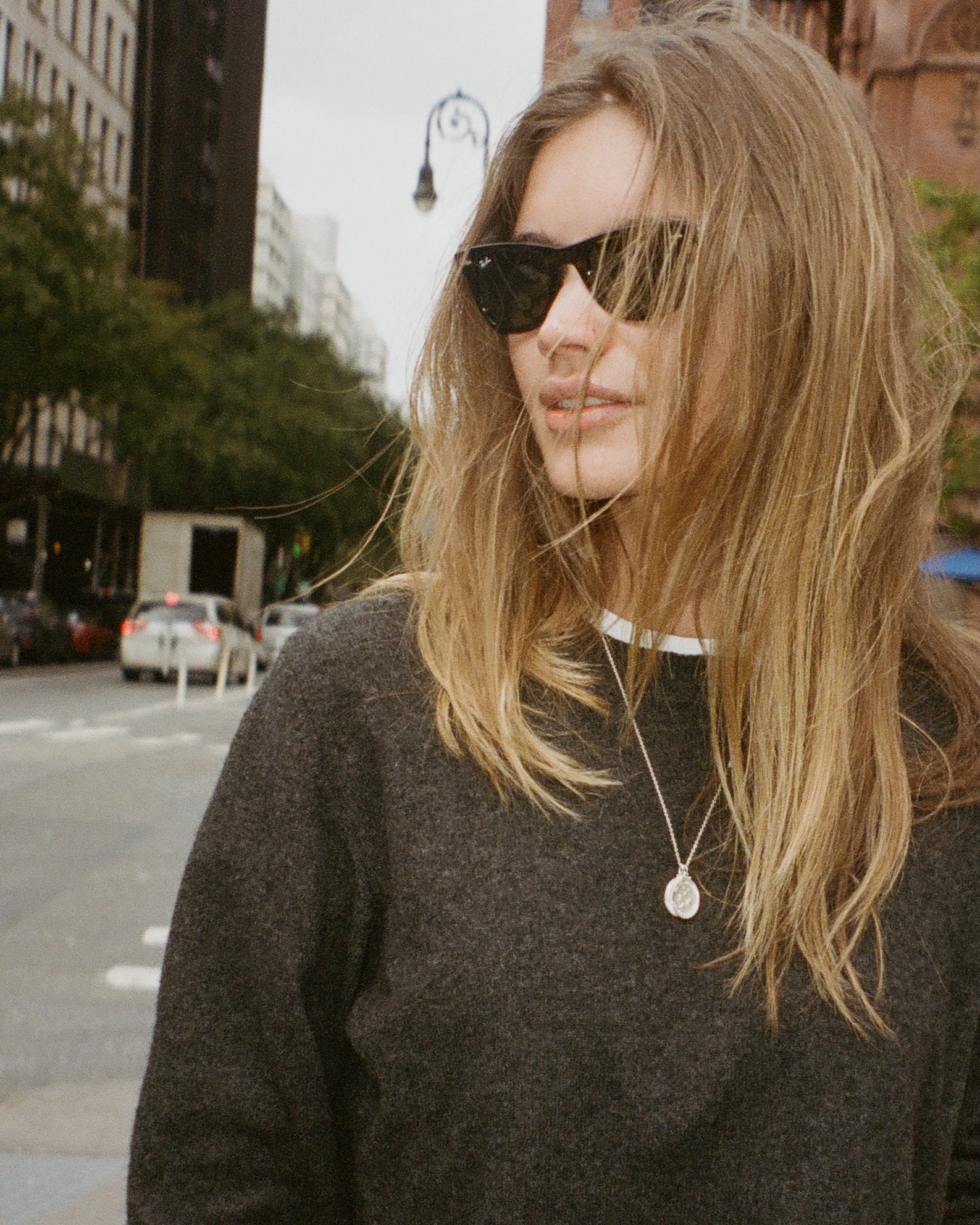 woman in a grey sweater wearing black ray ban sunglasses shown crossing a New York City street. She has on a love billy medallion necklace.