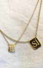 Two delicate gold chain necklaces with square initial pendants, one engraved with the letter 'B' and the other with 'S', displayed on a textured off-white fabric background.
